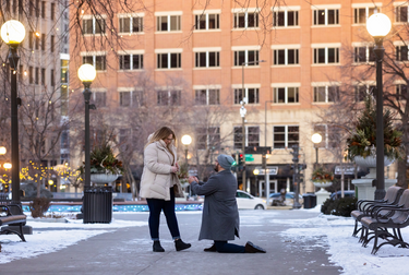 Man proposing to a woman in a snowy city park with buildings in the background.