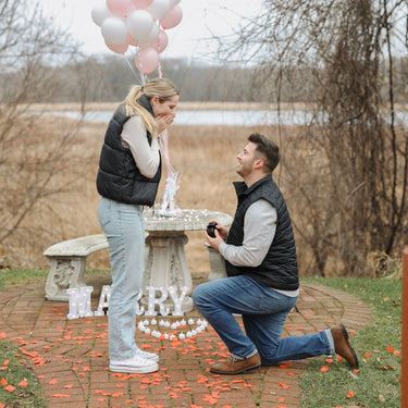 Man proposing to a woman with balloons and 'Will You' sign outdoors.